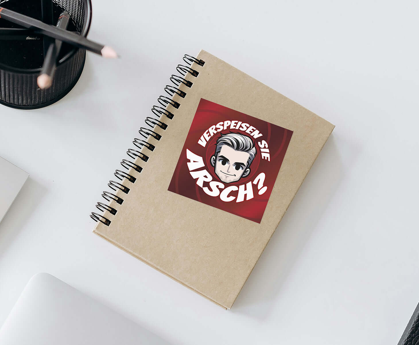 Kreatives Notizbuch mit humorvollem Aufdruck "VERSPEISEN SIE ARSCH?" auf einem Tisch mit Stiften.